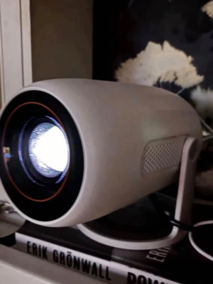Lenovo Xiaoxin 100M Projektor Full HD - Modern vit projektor med rundad form och svart/orange detalj runt linsen. Står på ett stabilt stativ. Perfekt för att streama film eller spela spel på stor skärm. Ansluts med strömkabel och har en stilren design. Inbyggt Android system för appar och HDMI för att koppla in AppleTV/Chromcast eller spelkonsoll. Upp till 120 tums bild. 1080p full HD väldigt kapabel projektor för pengarna.