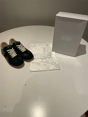 Maison Margiela svarta sneakers - Maison Margiela sneakers i svart mocka och läder med vita kontrasterande snören och beige gummisula. Klassisk låg modell med stilren design och ikonisk logotyp på insidan. Kommer med originalpåse och box.