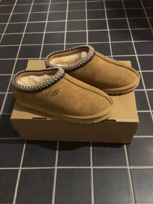 Bruna UGG tofflor i mocka - Säljer ett par klassiska bruna UGG tofflor i mocka med mjukt, vitt foder och dekorativt mönster runt öppningen. Tofflorna har en robust sula och UGG-loggan på sidan. Perfekta för att hålla fötterna varma hemma.