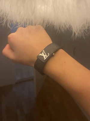 Louis Vuitton svart läderarmband - Snyggt svart armband från Louis Vuitton i präglat läder med det klassiska LV-märket i silverfärgad metall framtill. Armbandet har monogramdetaljer och ett stilrent spänne. Perfekt accessoar för dig som gillar exklusiv och trendig stil.