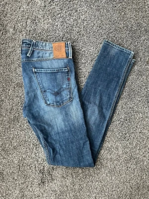 Blå smala jeans från Replay - Snygga blå jeans från Replay med smal passform och klassisk femficksdesign. Jeansen har en lätt tvättad look med slitningar och detaljer som läderpatch bak i midjan och röda sömmar. Perfekta för en trendig och avslappnad stil. Storlek 32/32 sitter mindre som 30/30