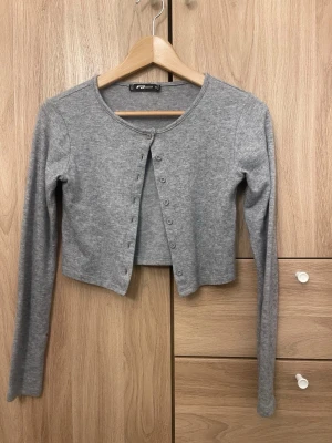 Grå cropped kofta  - Snygg grå cropped kofta med rund halsringning och långa ärmar. Koftan har knäppning framtill med små knappar och är tillverkad i ett mjukt, stretchigt material. Perfekt att styla över ett linne eller t-shirt.