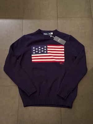 Mörkblå stickad tröja Ralph Lauren - Snygg mörkblå stickad tröja från Ralph Lauren med amerikansk flagga i rött, vitt och blått på bröstet. Tröjan har rund halsringning, ribbade muddar och nederkant samt RL-broderi. Perfekt för dig som gillar klassisk och preppy stil.