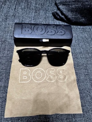 Svarta solglasögon från BOSS - Stilrena svarta solglasögon från BOSS med klassisk rektangulär form och diskret logga på skalmen. Glasögonen har svarta bågar i syntetmaterial och mörka linser. Perfekta för att lyfta din look under soliga dagar.