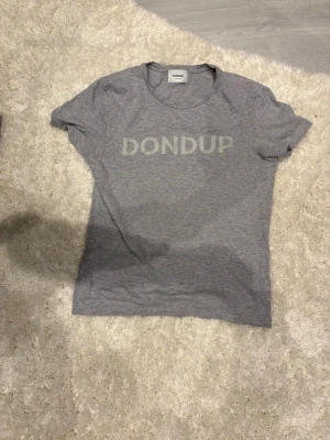 Grå t-shirt från Dondup - Snygg grå t-shirt från Dondup med tryckt logga vertikalt på framsidan. Klassisk rund hals och korta ärmar. Tillverkad i mjukt bomullsmaterial som känns skönt mot huden. Perfekt för en avslappnad och stilren look.
