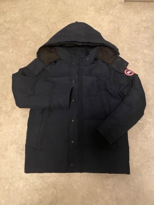 Mörkblå dunjacka från Canada Goose - Säljer en mörkblå dunjacka från Canada Goose med huva och klassisk logotyp på ärmen. Jackan är tjock, har tryckknappar framtill och är perfekt för kalla vinterdagar. Stilig och funktionell design som håller dig varm hela säsongen.