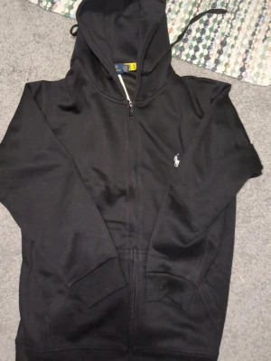 Svart zip hoodie från Polo Ralph Lauren - Snygg svart hoodie med dragkedja från Polo Ralph Lauren. Klassisk design som aldrig är ur modet,samt den ikoniska vita logon broderad på bröstet. Tillverkad i mjukt bomullsmaterial och har fickor framtill. Perfekt för en chill och stilren look. Priset är inte hugget i sten.