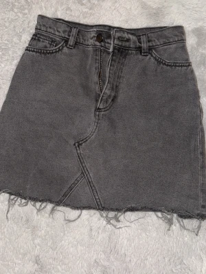 Grå jeanskjol med rå kant - Säljer en grå kort jeanskjol med rå, fransig kant och cool asymmetrisk söm fram. Kjolen har bälteshällor, knapp och dragkedja samt två bakfickor. Perfekt för dig som gillar en avslappnad och trendig stil.