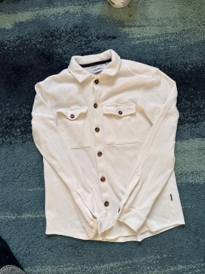 Vit skjorta från Strömberg - Klassisk vit skjorta från Strömberg i bomull med lång ärm och bruna knappar. Skjortan har två bröstfickor med lock och en stilren krage. Perfekt för dig som gillar enkel och clean stil.