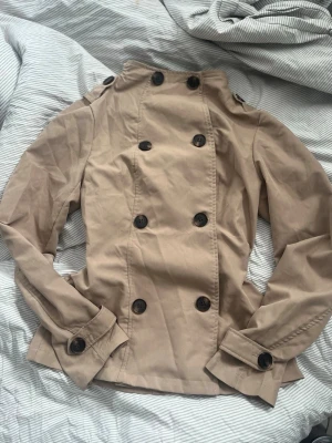 Beige trenchcoat från Muchica - Stilren beige trenchcoat från Muchica med dubbelknäppning och högt krage. Den är varm och mysig och bara använd en gång. Skriv till mig om ni vill ha mer bilder på jackan💕