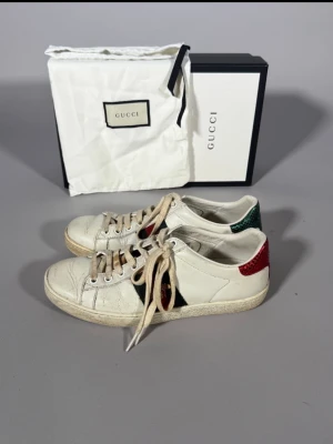 Gucci vita sneakers med röd/grön detalj - Snygga vita sneakers från Gucci i läder med klassiska gröna och röda ränder på sidan. Baktill har ena skon en röd detalj och den andra en grön, vilket ger en unik touch. Låga i modellen och med vita snören. Perfekta för dig som gillar exklusiv streetstyle.