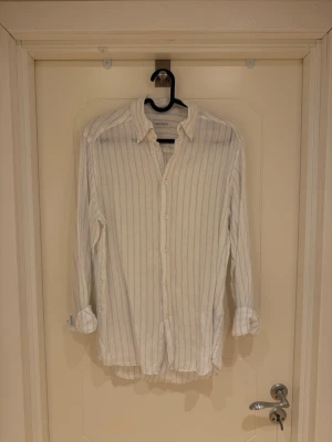 Vit randig linneskjorta These Glory Days - Klassisk vit skjorta från These Glory Days med tunna blå ränder. Slim fit-modell med långa ärmar och knappar framtill. Tillverkad i linne som känns skön mot huden. Perfekt för en clean och stilren look.                                       Modellen är 185cm lång och 82kg.