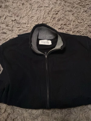 Svart ziptröja från Pier One - Snygg svart ziptröja från Pier One med hög krage och grå insida. Tröjan har långa ärmar, två fickor framtill och ribbade muddar. Perfekt för en avslappnad och stilren look.