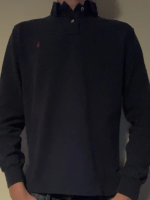 Mörkblå långärmad skjorta Polo Ralph Lauren - Klassisk mörkblå långärmad skjorta från Polo Ralph Lauren med liten röd logga broderad på bröstet. Skjortan har krage, knappar framtill och är tillverkad i mjuk bomull. Perfekt för en stilren och avslappnad look. Storlek S-M mer åt S hållet 