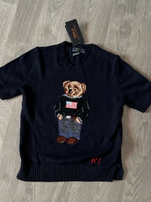 Mörkblå stickad tröja Polo Bear - Mörkblå stickad tröja från Polo Ralph Lauren med ikoniskt Polo Bear-motiv framtill. Tröjan har korta ärmar, ribbade muddar och rund halsringning. Materialet är mjukt och bekvämt, perfekt för en avslappnad men stilren look.