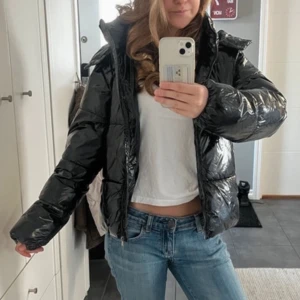Svart glansig pufferjacka med huva - Säljer en svart, glansig pufferjacka med avtagbar huva och dragkedja framtill. Perfekt för kalla dagar och riktigt varm. Inga tecken på användning, ser helt ny ut. Nypris 799kr.