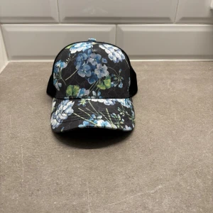 Svart keps med blått blommönster GG - Snygg svart keps med blått och vitt blommönster framtill. Kepsen har böjd skärm och mesh på sidorna och bak för extra ventilation. Perfekt för dig som vill sticka ut med en unik accessoar. Justerbar baktill för bästa passform. Tar emot bud