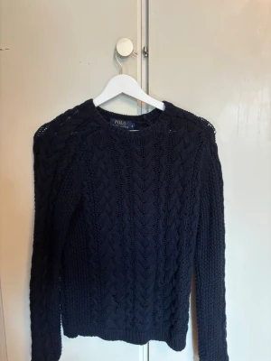 Mörkblå kabelstickad tröja Polo Ralph Lauren - Snygg mörkblå kabelstickad tröja från Polo Ralph Lauren. Tröjan har rund halsringning, långa ärmar och klassiskt flätmönster. Perfekt för dig som gillar tidlös och stilren design. Materialet känns mjukt och håller värmen bra.