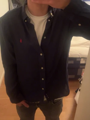 Mörkblå skjorta från Ralph Lauren - Klassisk mörkblå skjorta från Ralph Lauren med röd broderad logga på bröstet. Mycket bra skick, 8/10. Priset kan diskuteras! Hör av er ifall ni har några frågor!!