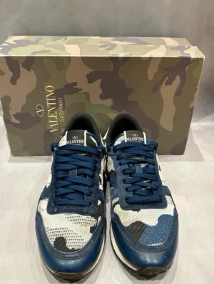 Valentino Garavani Rockrunners blå camo sneakers - Snygga sneakers från Valentino Garavani Rockrunners med blå, vita och svarta camo-detaljer. Storlek 40, men passar 40-41. Ovandelen är i mesh och mocka, med vita och svarta inslag. Skorna är i bra skick. Perfekta för dig som vill sticka ut och ha ett par lite skönare dojjor. Hör av er vid frågor och funderingar!