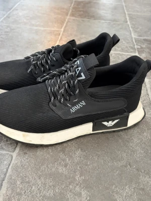 Svarta sneakers från Armani - Behöver torkas av. Därav billigt pris. Men hela och fina! Skit snygga. Stilrena svarta sneakers från Armani med vita detaljer och logga på sidan. Skorna har svart ovandel i textil, vita och svarta snören samt vit sula med svart parti och EA7-logga. Perfekta för dig som gillar en clean och modern look.