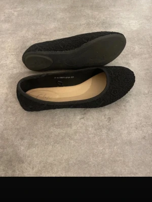 Svarta spetsballerinaskor - Säljer ett par svarta ballerinaskor med spetsdetaljer och rundad tå. Skorna har en tunn sula och en klassisk, enkel design som passar till många olika outfits. Insidan är beige och materialet på ovandelen är täckt av ett blommigt spetsmönster.