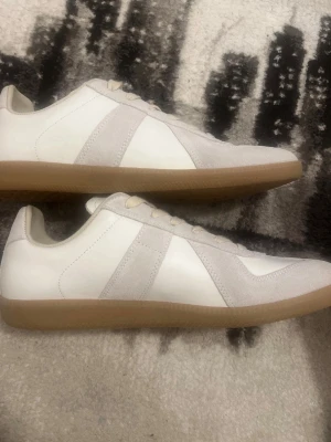 Maison margiela  - Snygga sneakers i vitt och ljusgrått med ovandel i mocka och skinn. Klassisk design med snörning och platt sula i gummi. Perfekta för dig som gillar stilrena och tidlösa sneakers. Märket Maison Margiela väldigt fina o sköna skor passar perfekt inför kommande årstider våren och sommaren väldigt stiliga och passar med allt. PRISET KAN DISKUTERAS:) Kvitto finns!!