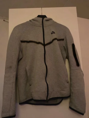 Grå hoodie med dragkedja från Nike - Snygg grå hoodie från Nike med hel dragkedja framtill och svart Nike-logga på bröstet. Hoodien har svarta detaljer längs dragkedjan och ärmarna, samt en ficka på ena ärmen. Tillverkad i mjukt sweatshirtmaterial och har en skön huva.