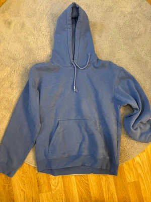 Blå hoodie med magficka - En enkel blå hoodie med huva och dragsko. Hoodien har en stor magficka framtill och ribbade muddar vid ärmslut och nederkant. Perfekt för en avslappnad och chill stil. Märke och material är okänt. Ena snöret är lite trasigt annars inga fel
