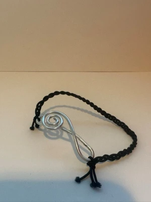 Armband med silverfärgad G-klav - Armbandet är gjort av flätad svart tråd, fäst till tråden är en silverfärgad G-klav. G-klaven är gjord på silverfärgad metalltråd som är böjd för hand. Perfekt för dig som vill ha ett kreativt smycke. 