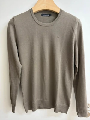 Beige stickad tröja från J.Lindeberg i merinoull - Snygg beige stickad tröja från J.Lindeberg med rund halsringning och diskret broderad logga på bröstet. Tröjan har långa ärmar och ribbade muddar vid ärmslut och nederkant. Perfekt för en stilren och enkel look.