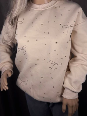 Beige sweatshirt med pärlor och rosetter - Supersöt beige sweatshirt med små pärlor och broderade rosetter framtill. Tröjan har rund halsringning, långa ärmar och mjukt material som känns skönt mot huden. Perfekt för dig som gillar gulliga detaljer och en clean look.