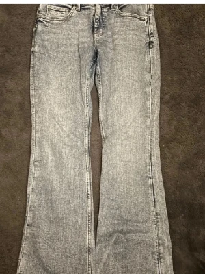 Grå bootcut jeans med tvättad look - Säljer ett par blå bootcut jeans med snygg tvättad look och klassiska fem fickor. Jeansen har låg midja och utsvängda ben, perfekta till sneakers eller boots. ALDRIG ANVÄNDA!