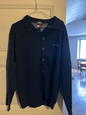Mörkblå cardigan från Tommy Hilfiger - Mörkblå cardigan med knappar framtill och klassisk krage från Tommy Hilfiger. Stort broderat logotyp-tryck i vitt och rött på ryggen samt diskret logga på bröstet. Långärmad och i mjukt stickat material, perfekt för en stilren look. Vintage köpt i Paris förra året