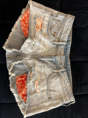 Ljusa denimshorts med slitningar - Säljer ett par ljusa denimshorts med råa kanter och slitna detaljer framtill. Shortsen har klassisk femficksdesign och coola röda bandana-detaljer i fickorna. Perfekta för sommaren och ger en avslappnad streetstyle-look.