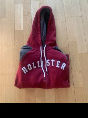 Röd Hollister hoodie med dragkedja - Snygg röd hoodie från Hollister med vit logga på bröstet. Hoodien har dragkedja, vita snören i huvan och gråa partier på axlarna samt vita detaljer på ärmarna. Mjuk och skön bomullsblandning, perfekt för chill dagar.
