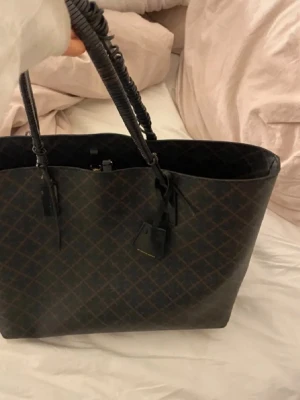 Brun shoppingväska By Malene Birger - Stilren shoppingväska från By Malene Birger i mörkbrunt konstläder med rutigt mönster och svarta handtag. Väskan är rymlig och har en klassisk design med diskret logga och detaljer i svart. Perfekt för dig som gillar trendiga och praktiska accessoarer.