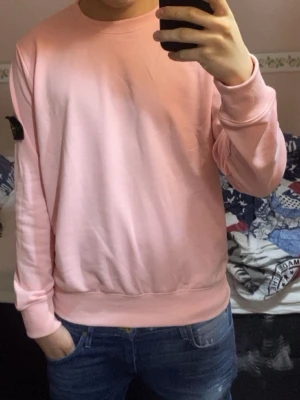 Ljusrosa sweatshirt från Stone Island - Ljusrosa sweatshirt från Stone Island med klassisk rund hals och ribbade muddar. Tröjan har den ikoniska svarta patchen med gul och grön kompasslogga på vänster ärm. Tillverkad i mjuk bomull för en skön och avslappnad look.