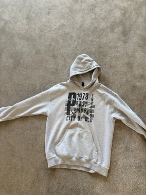 Grå hoodie med tryck London 1973 - Säljer en grå hoodie med svart tryck framtill där det står '1973 East London City of Old' och motiv av Union Jack. Hoodien har huva och en stor magficka. Perfekt för dig som gillar streetwear och vill ha en avslappnad stil.