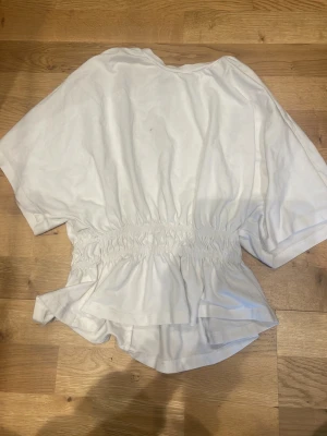 Vit topp💕🤍 - Säljer en vit topp med korta ärmar och resår i midjan som ger en snygg klang. Toppen har en lös och luftig design, perfekt för varmare dagar. Materialet känns mjukt och bekvämt mot huden. Jag har tyvär fått en mascara fläck på tröjan som inte går bort som man ser på bilderna. Har använt kanske 3 gånger. 💝😊💕