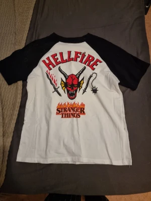 Stranger Things Hellfire Club t-shirt - Cool t-shirt med Hellfire Club-tryck från Stranger Things. Vit bas med svarta korta ärmar och färgglad grafik på ryggen. Perfekt för fans av serien som vill sticka ut med en unik och snygg tee. Litet hål i sidan. XXS men den är stor, och passar nog ända upp till M. Aldrig använt 