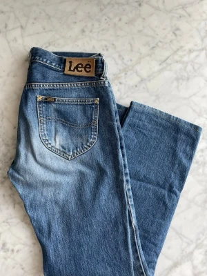 Blå raka jeans från Lee - Säljer ett par klassiska blå jeans från Lee med rak passform och fem fickor. Jeansen har en snygg tvättad look och Lee-logga på bakfickan. 100% bomull. Lite slitet framtill mellan låren.