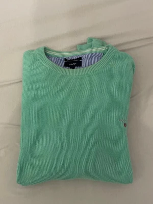 Mintgrön stickad tröja från Gant - Snygg mintgrön stickad tröja från Gant med rund halsringning och diskret logga broderad på bröstet. Tröjan har ribbade muddar vid ärmslut och nederkant. Perfekt för dig som gillar stilrena och fräscha plagg i garderoben.