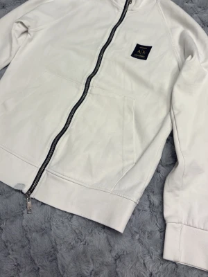 Vit ziptröja från Armani Exchange - Snygg vit ziptröja från Armani Exchange med svart logopatch på bröstet. Tröjan har hel dragkedja, två stora fickor framtill och ribbade muddar vid ärmslut och nederkant. Perfekt för en clean och stilren look. Finns lite hål på den. Kolla noga på bilderna. 