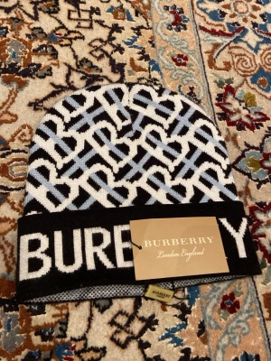 Burberry svartvit mönstrad mössa - Säljer en snygg svartvit mössa från Burberry med grafiskt monogrammönster och bred vikt kant där BURBERRY står i stora bokstäver. Mössan är stickad och har inslag av ljusblått i mönstret. Perfekt accessoar för dig som gillar streetstyle och designerplagg.