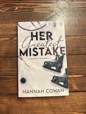 Her Greatest Mistake av Hannah Cowan - En romantisk roman på engelska med hockeytema, skriven av Hannah Cowan. Omslaget är ljust med svarta skridskor, puckar och klubba, vilket ger en sportig känsla. Perfekt för dig som gillar kärlekshistorier med sportinslag.