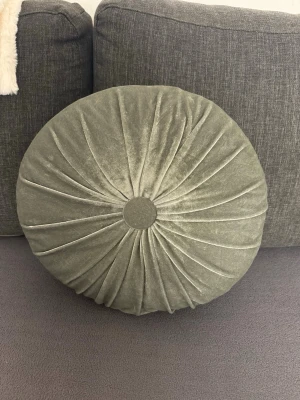 Rund grå sammetskudde - Säljer en rund kudde i mjuk grön sammet med dekorativa veck och en knapp i mitten. Kudden har en lyxig känsla och passar perfekt som inredningsdetalj i soffan eller på sängen.