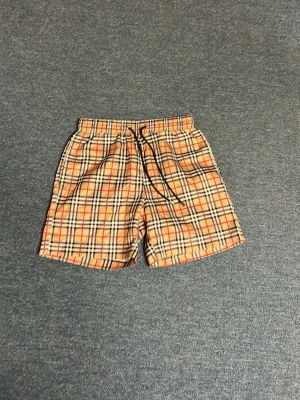 Rutiga shorts med snörning - Snygga shorts med klassiskt rutig mönster i beige, svart och rött. De har elastisk midja med svart snörning och är perfekta för sommaren. Materialet känns lätt och luftigt, vilket gör dem sköna att bära under varma dagar. (Badshorts)