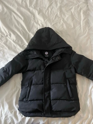 Svart dunjacka från Canada Goose - Säljer en svart dunjacka från Canada Goose med huva och dragkedja. Jackan har stora fickor framtill och klassisk patch på ärmen. Perfekt för kalla vinterdagar och riktigt varm tack vare dunfyllningen. Storlek S-M