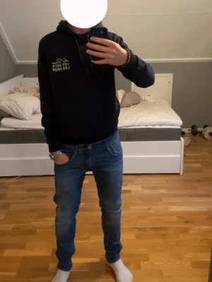 Mörkblå hoodie från Les Deux - Snygg mörkblå hoodie från Les Deux med vit logga tryckt på bröstet. Klassisk modell med huva och dragsnören samt en stor magficka. Tillverkad i mjukt material som passar perfekt till jeans eller joggers. Ribbade muddar vid ärmslut och nederkant. Passar dig från ca 170-180 cm
